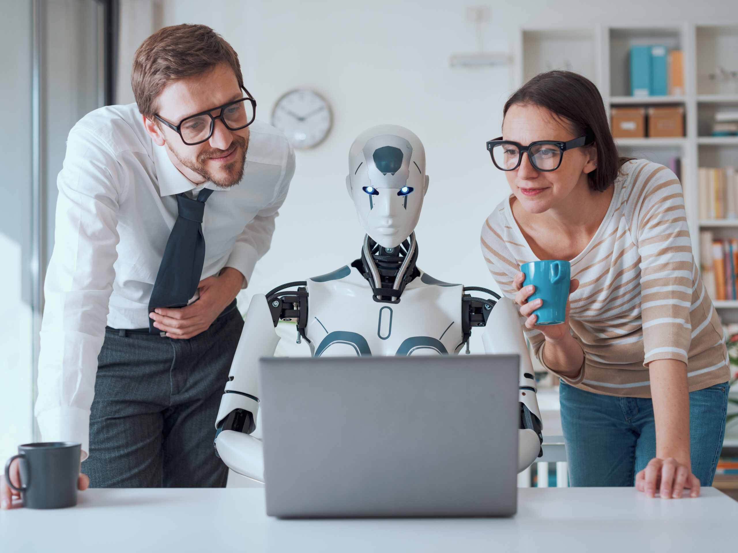 A man and a woman with glasses stand on either side of a robot or android working at a laptop