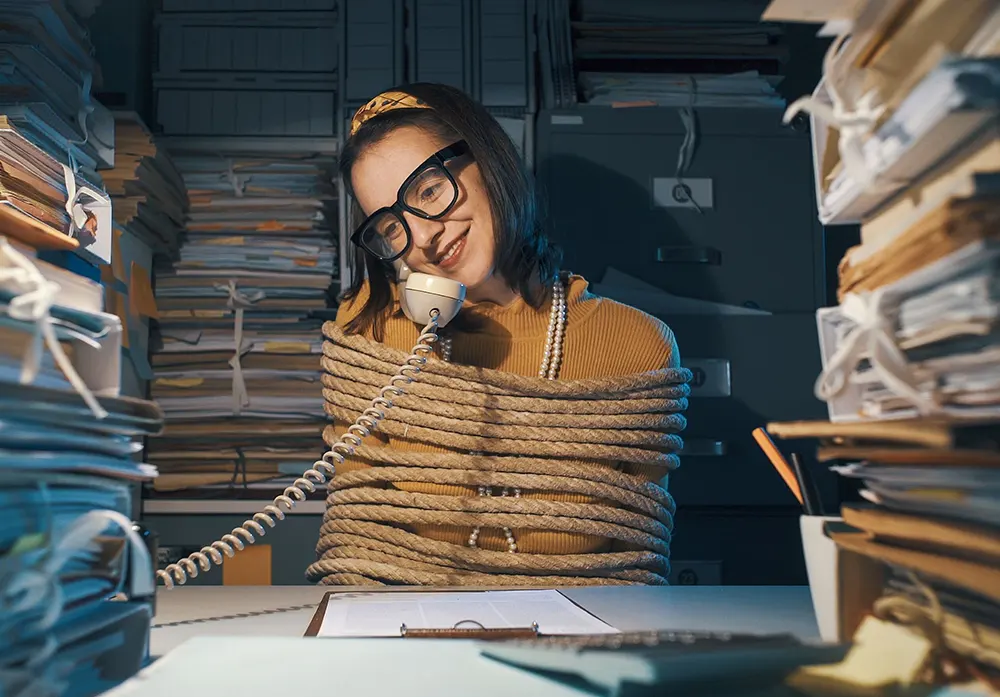 A smiling woman has a phone conversation while tied up at her desk with rope