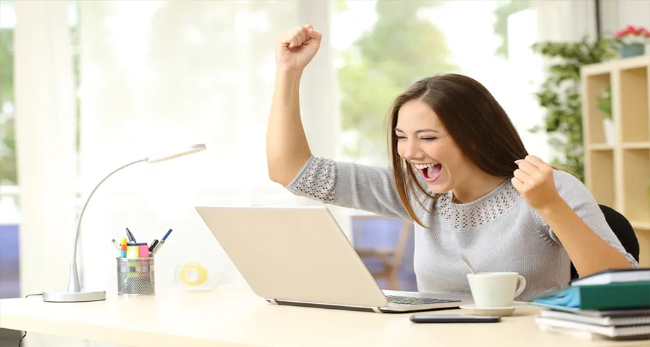 A happy woman in front of a laptop raises her fists in the air