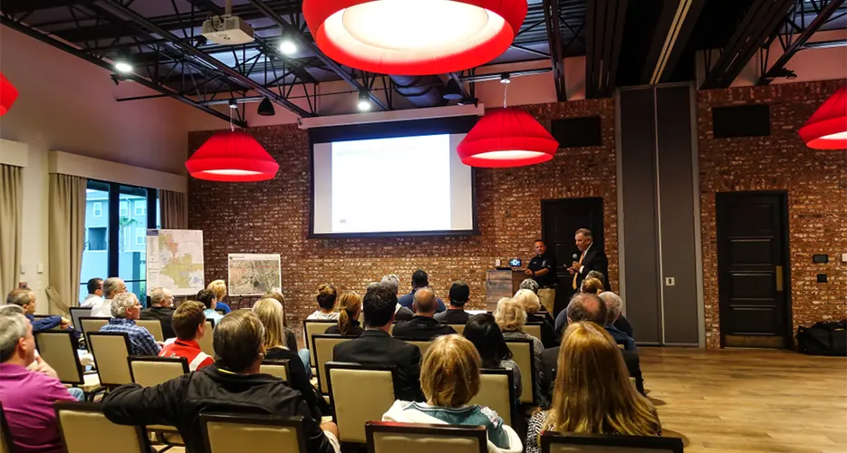 A man in a business suit speaks into a microphone in front of a group of seated people