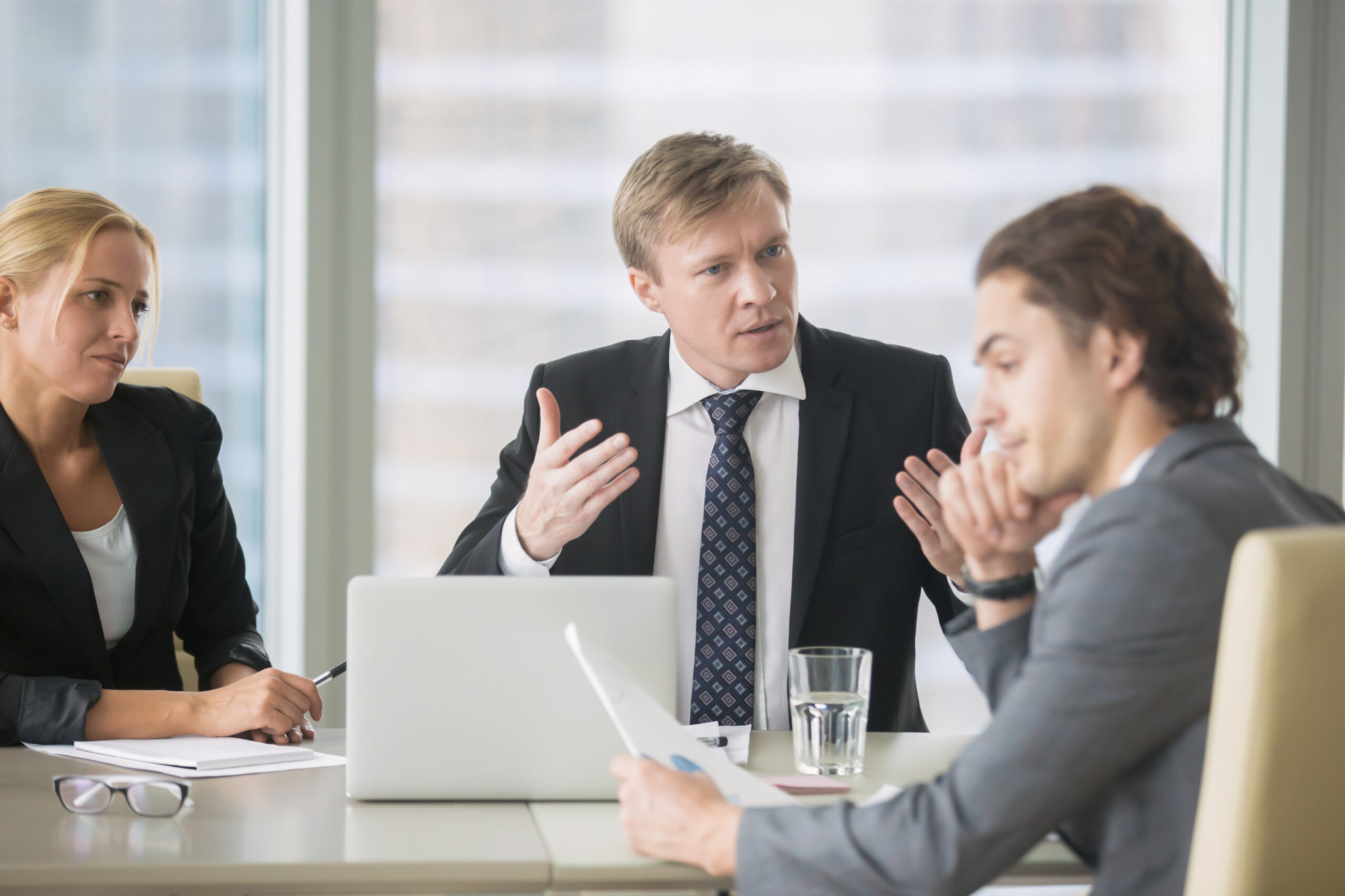 A businessman in a suit and tie reprimands an intern or young male office worker in a suit while a blonde woman looks on