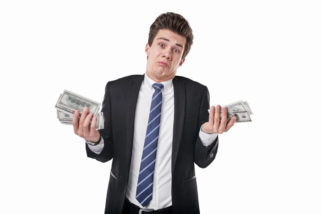 A man in a suit and tie holding two stacks of dollar bills shrugs.