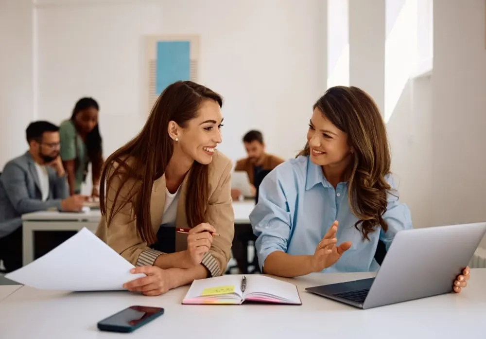 Two women smile at each other in front of papers and a laptop while collaborating on a creative work project.