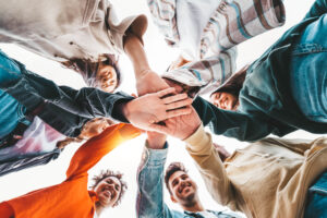 A group of young casually dressed students or professionals stand in a circle and stack a hand on top of each other's hand.