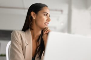A thoughtful professional woman, part of the Māori tribe, the indigenous Polynesian people of New Zealand (Aotearoa), looks out a window while sitting at a desk with a laptop.