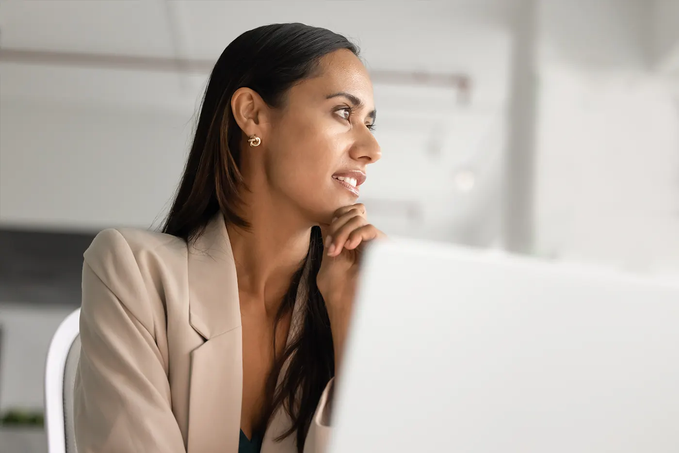 A thoughtful professional woman, part of the Māori tribe, the indigenous Polynesian people of New Zealand (Aotearoa), looks out a window while sitting at a desk with a laptop.